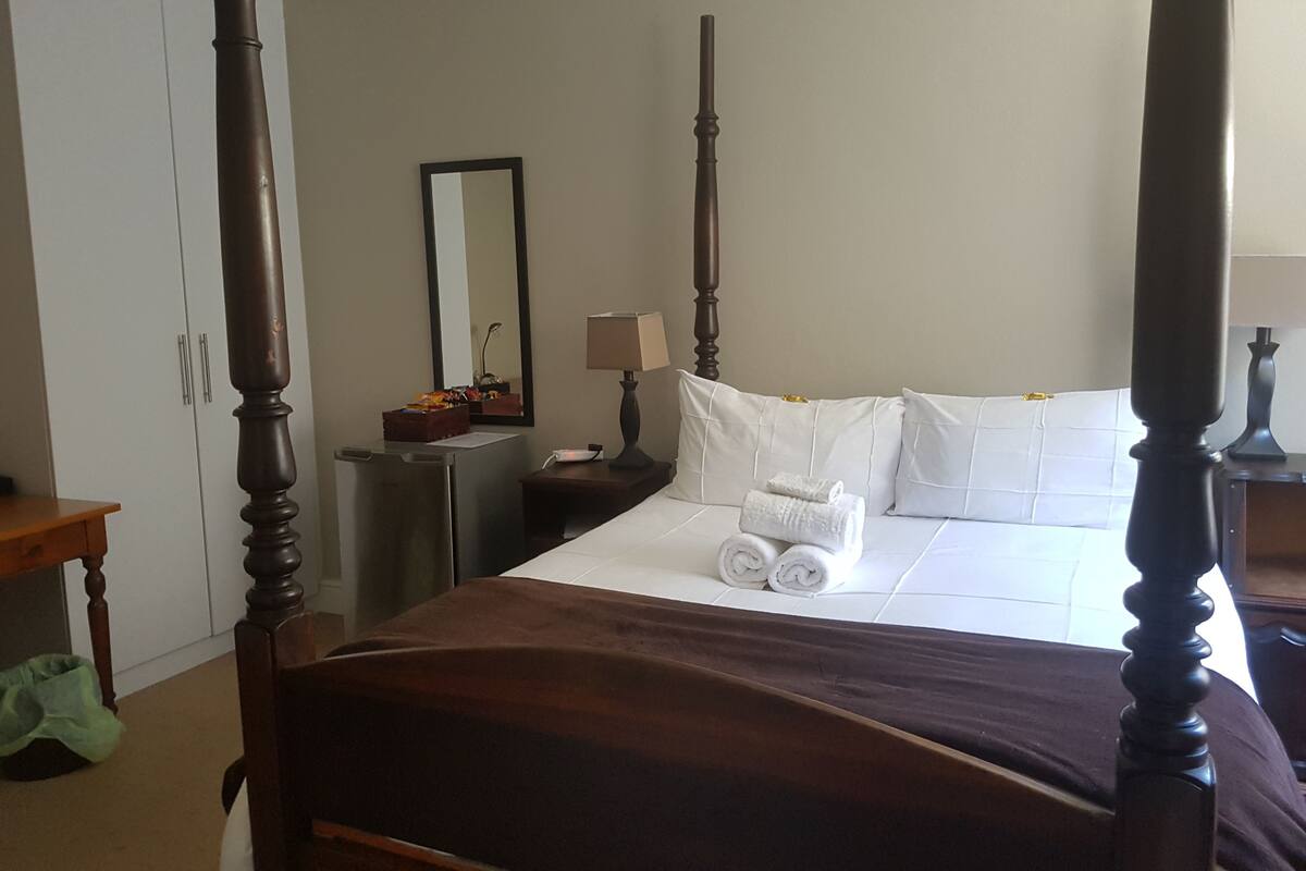 Uma cama de dossel com roupa de cama branca, uma mesinha de cabeceira com abajur e um espelho na parede.
