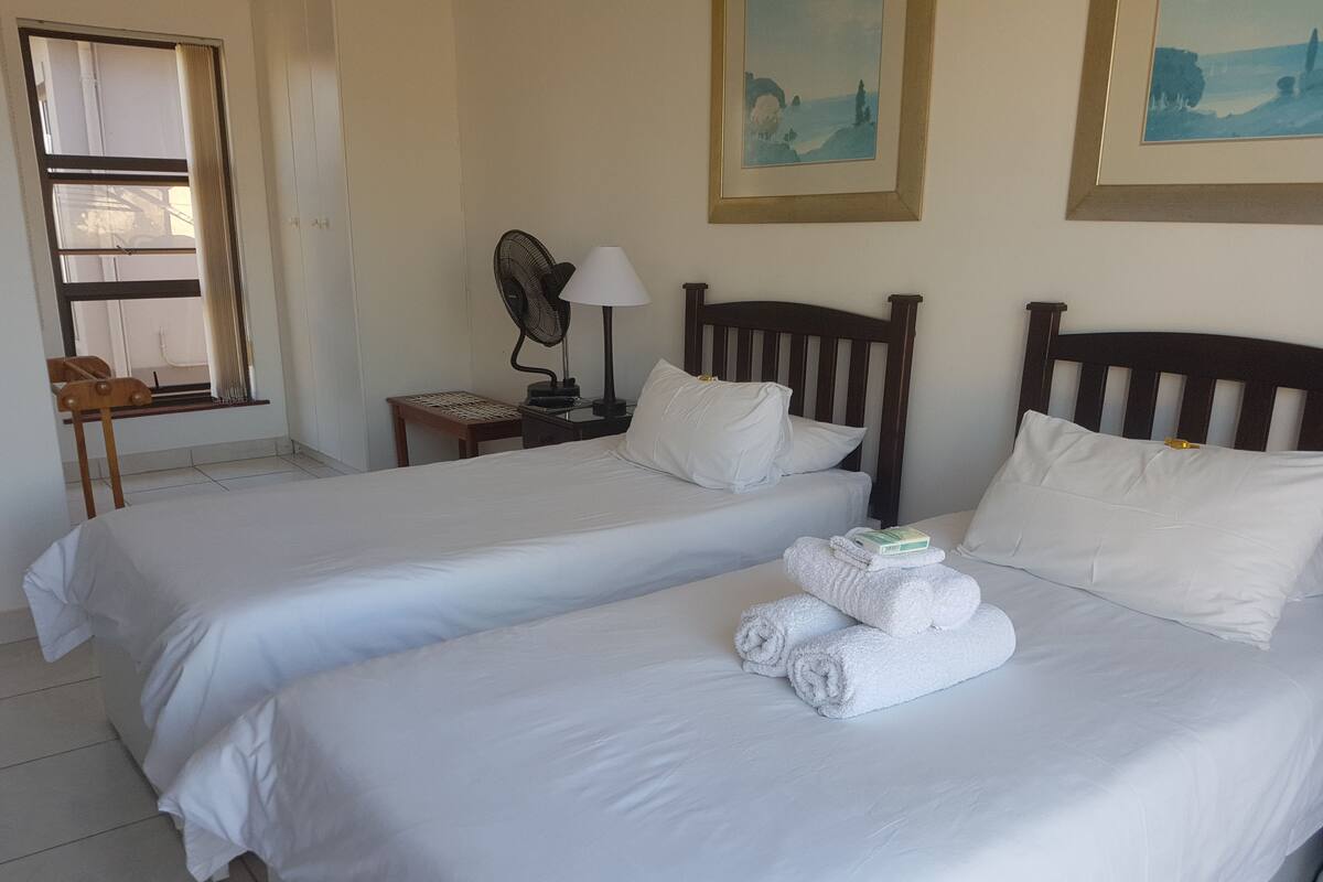 Quarto de hotel com duas camas de solteiro, mesa de cabeceira com abajur, ventoinha e janela com cortinas.