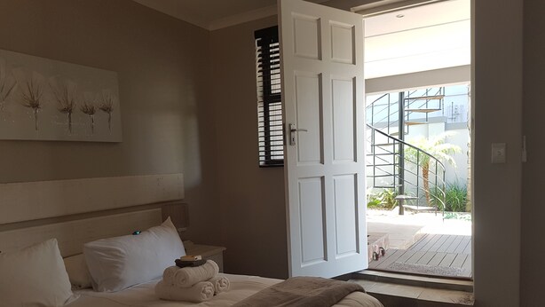 A bedroom with a bed, pillows, a bedside table, a nightstand, a painting, and a door leading to a balcony with a staircase and outdoor plants. - Melkbosch Guesthouse (Cape Town)