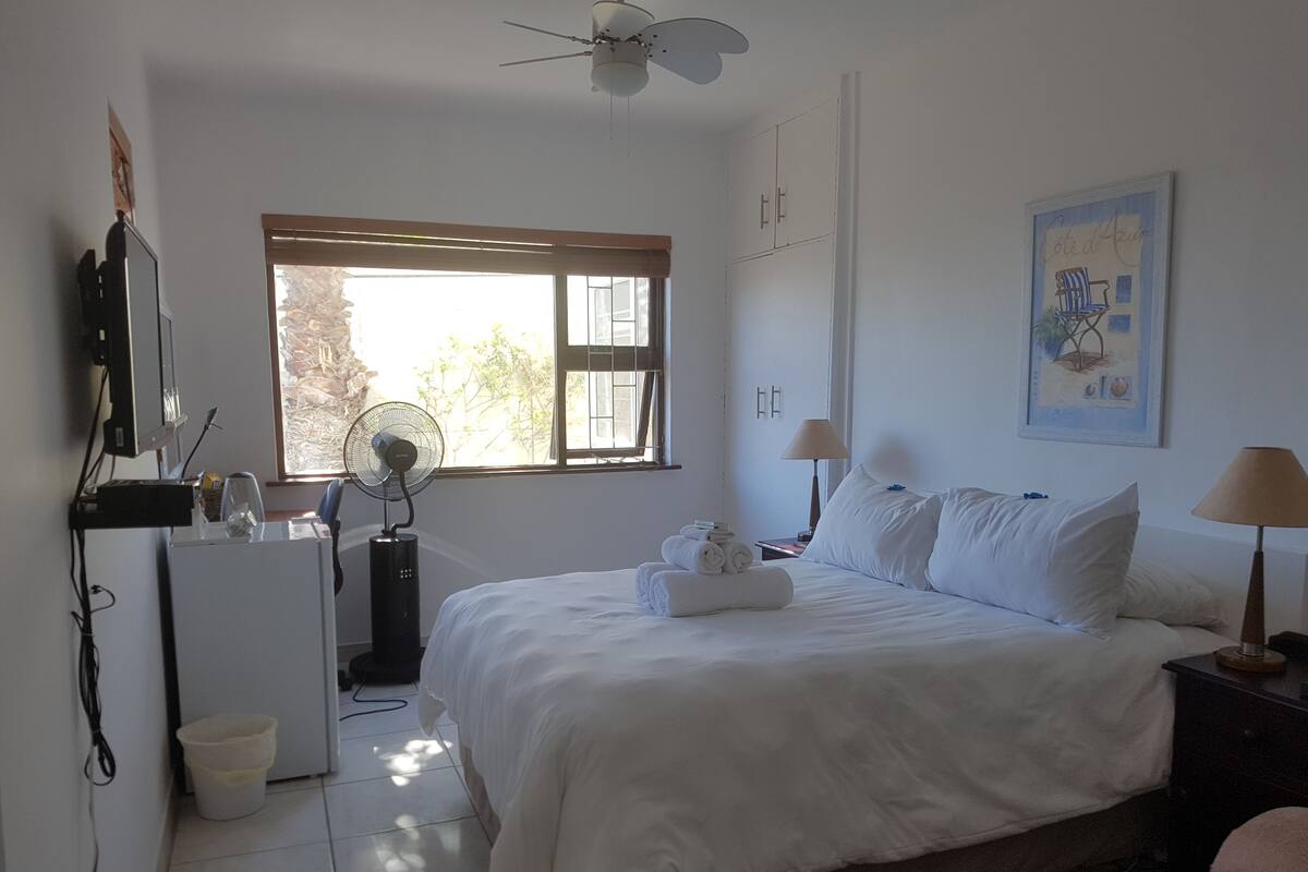 Quarto com cama, luminárias de cabeceira, escrivaninha com computador, ventilador e janela com persianas.
