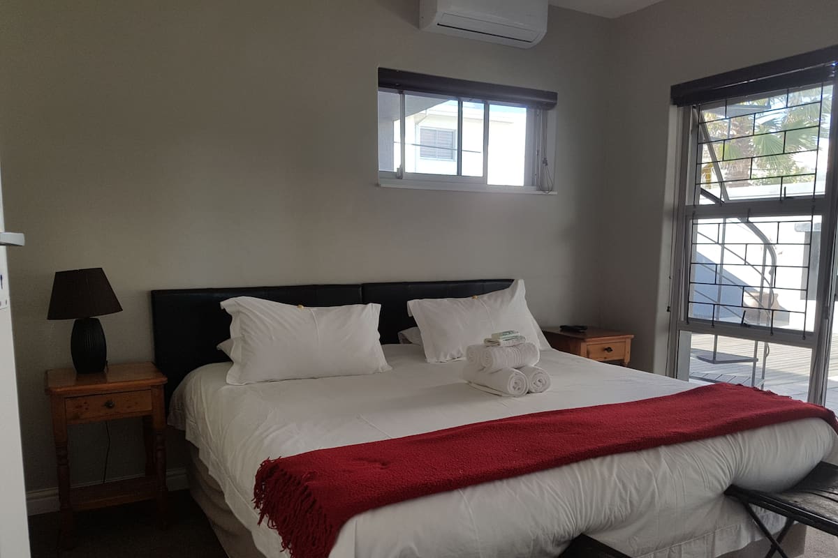 Uma cama bem arrumada com lençóis brancos e um cobertor vermelho, um abajur e uma janela com vista para palmeiras.