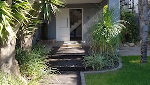 A modern two-story house with a wooden deck leading to a front door, surrounded by lush greenery and palm trees. - Melkbosch Guesthouse (Cape Town)