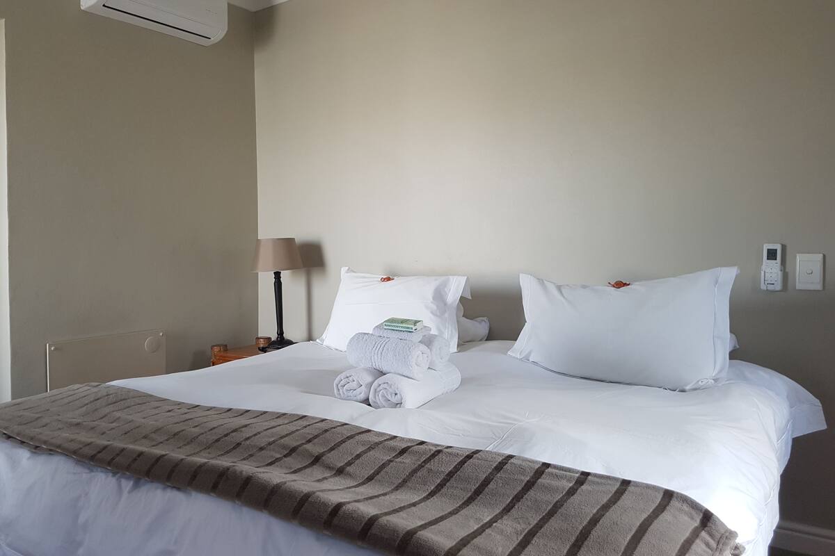 Quarto de hotel com cama, roupa de cama branca, manta listrada marrom, luminária e unidade de ar condicionado.