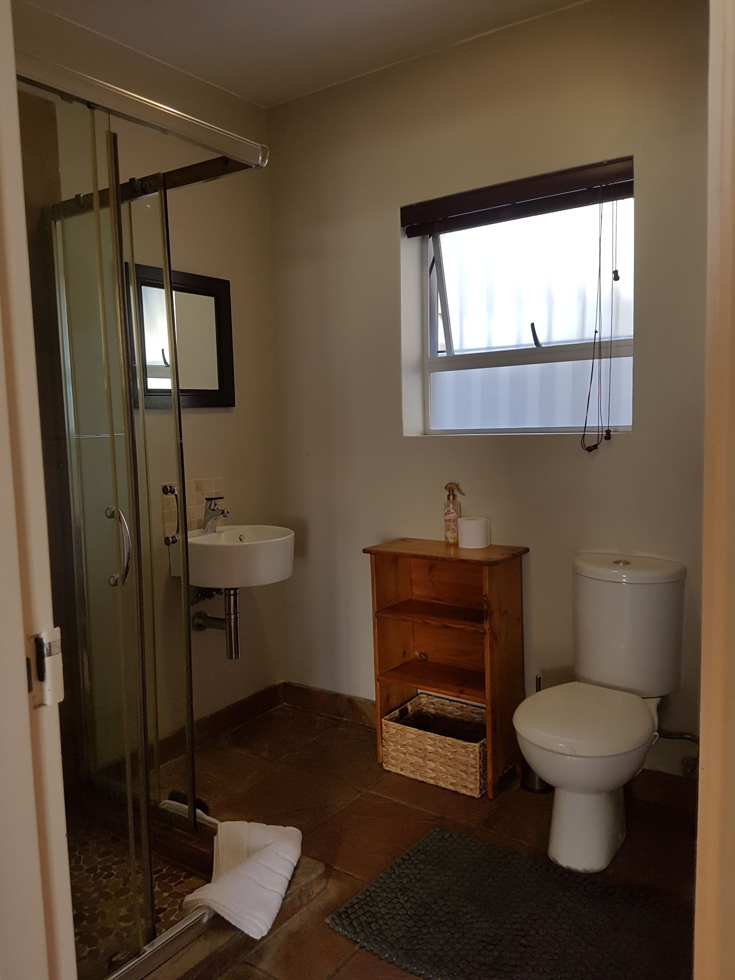 A bathroom with a glass shower enclosure, a sink, a toilet, a window with blinds, and a small wooden shelf with a basket.