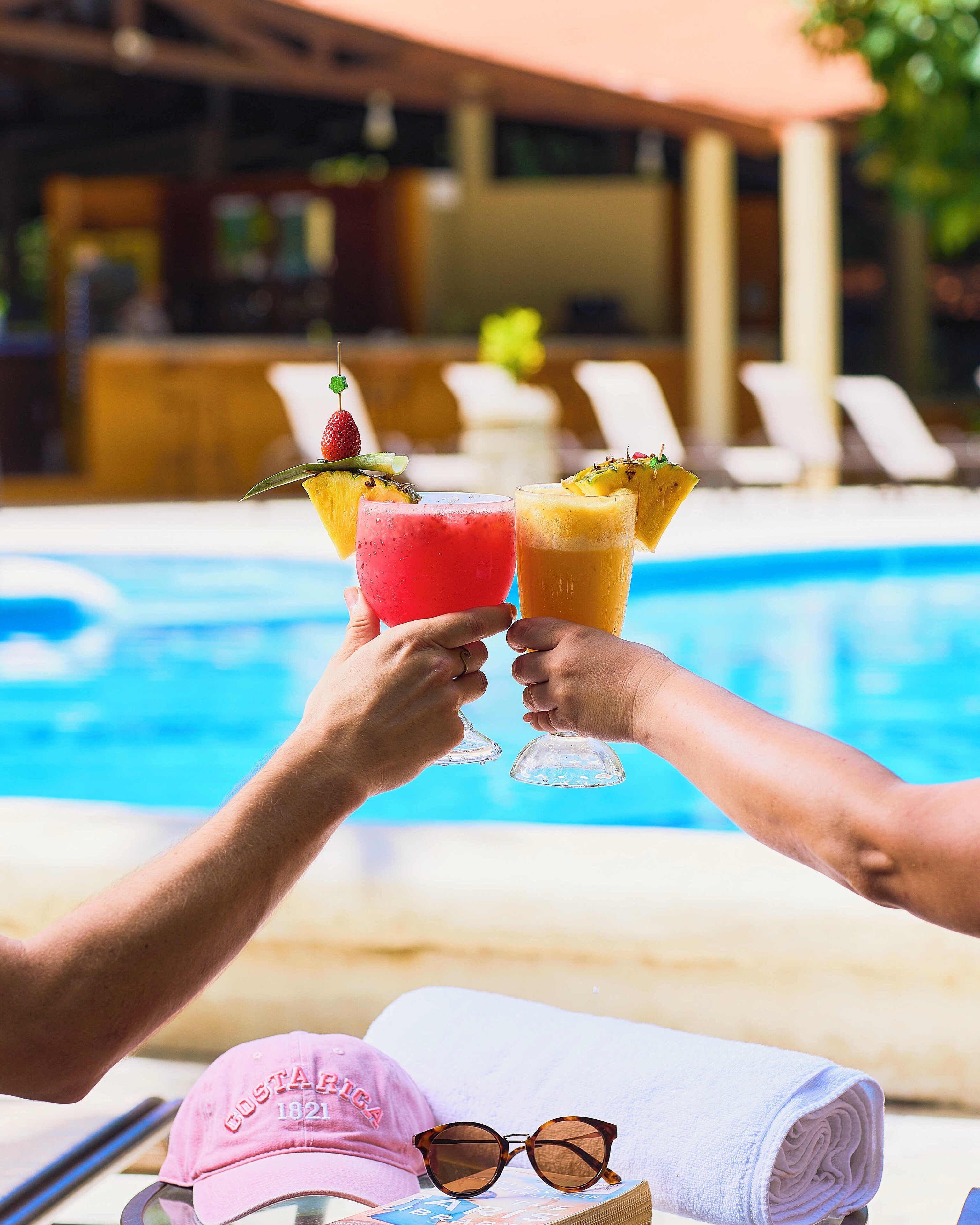 Two people toasting with drinks garnished with fruit, a pool, and a towel with sunglasses and a cap on the table.
