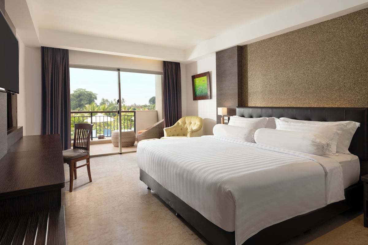 A hotel room with a large bed, a desk, a chair, a balcony with a view of greenery, and a painting on the wall.