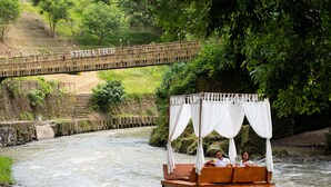 A wooden boat with a canopy and two people on it, floating on a river with a bridge in the background.