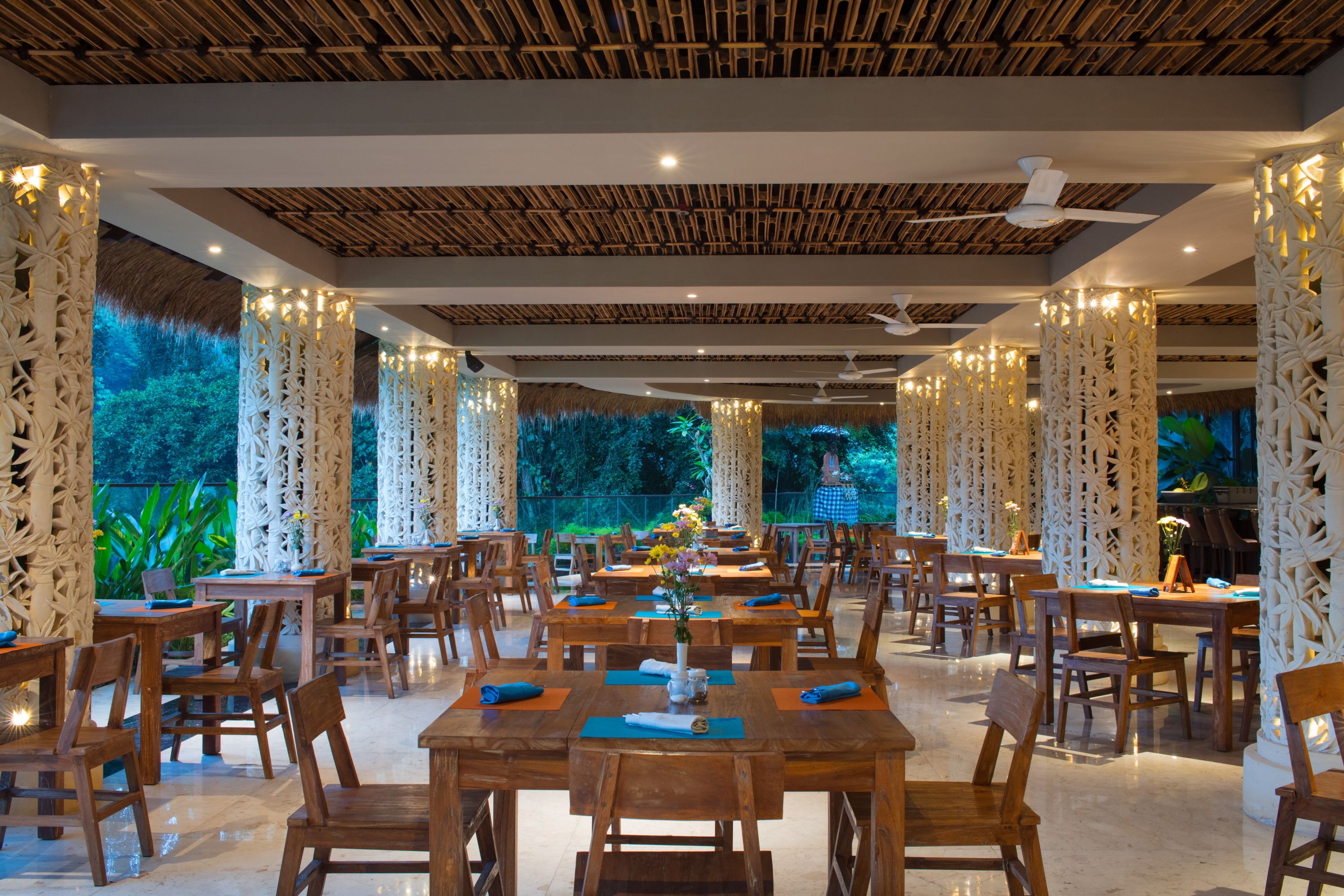 Un comedor amplio con mesas y sillas de madera, pilares blancos decorativos y vistas al exterior con vegetación.