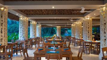 Un comedor amplio con mesas y sillas de madera, pilares blancos decorativos y vistas al exterior con vegetación.
