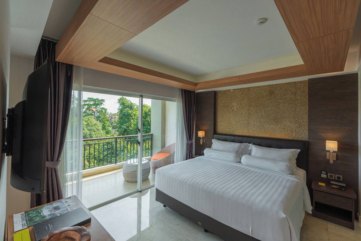 A modern hotel room with a large bed, a flat-screen TV, a balcony with a view of greenery, and a wooden ceiling.