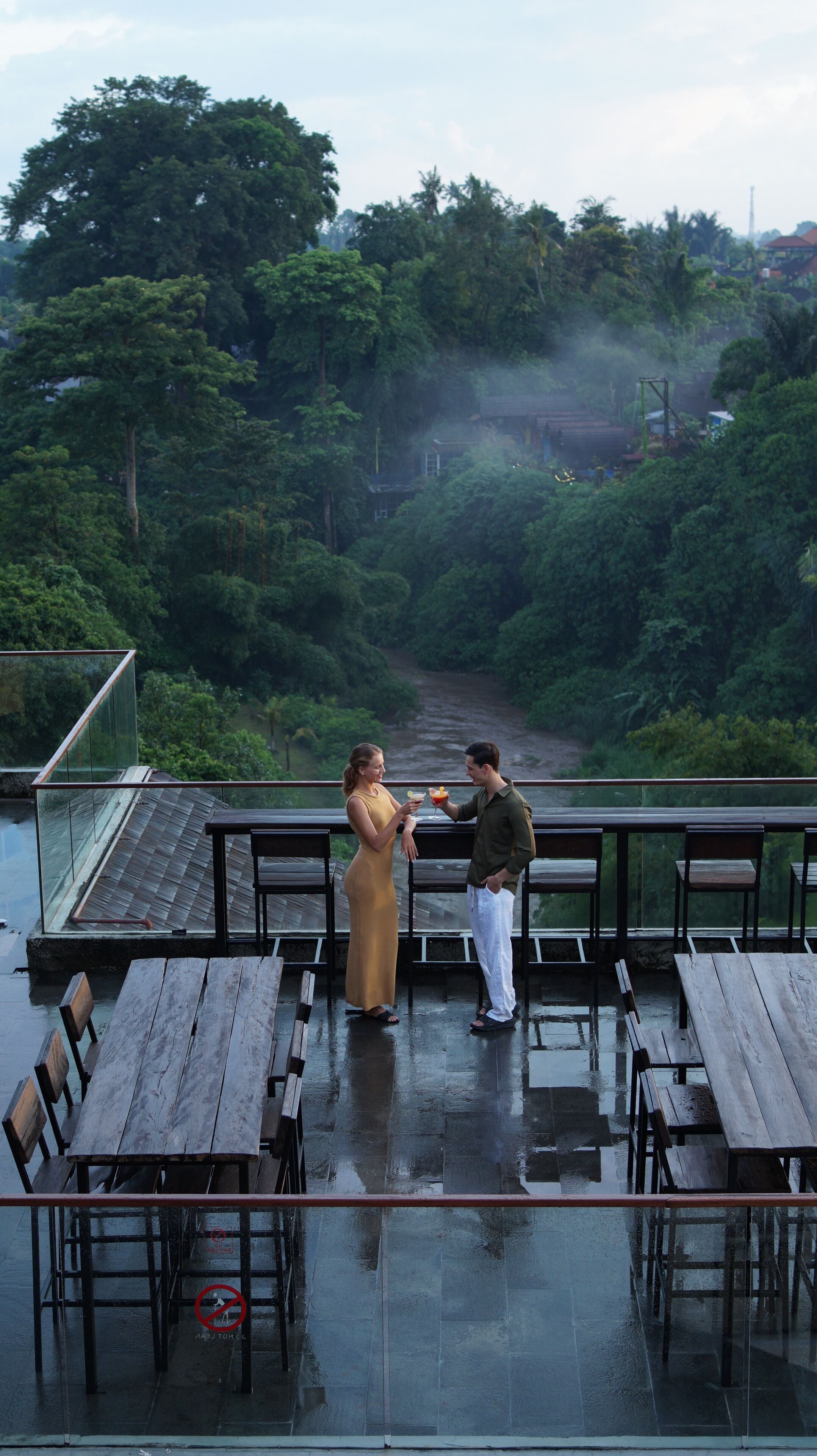 A couple standing on a wet wooden deck with a view of a river and dense greenery in the background.