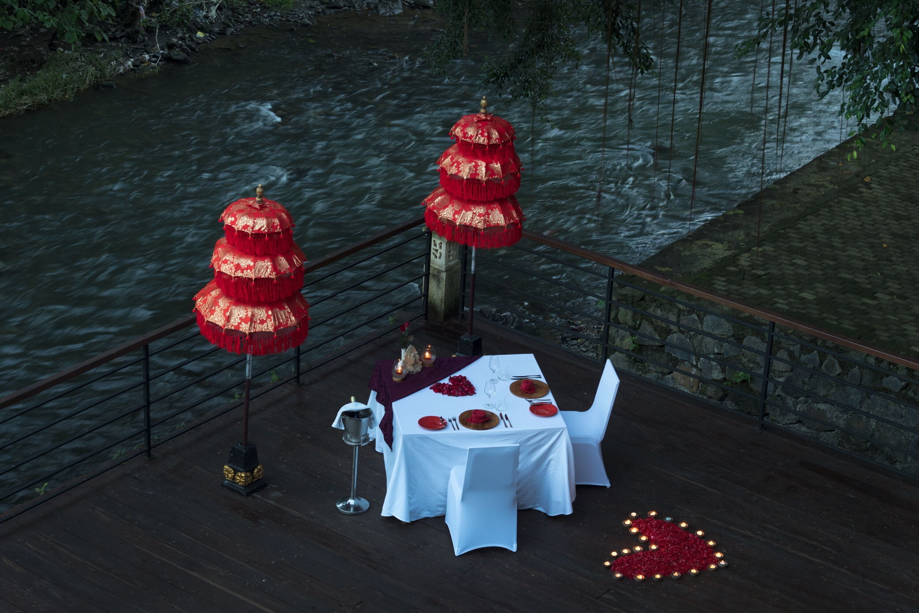 Una mesa de comedor decorada con manteles blancos, sombrillas rojas y doradas, y un arreglo floral rojo con forma de corazón.