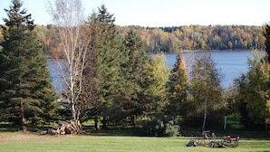 Blick auf einen See mit einer Wiese, Bäumen und einem Holzstapel.