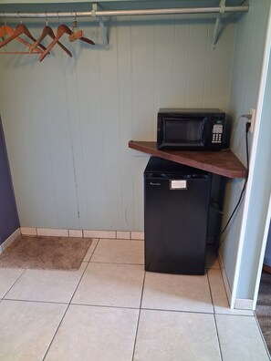 A compact kitchen with a microwave on a wooden shelf, a small refrigerator, and a tiled floor.