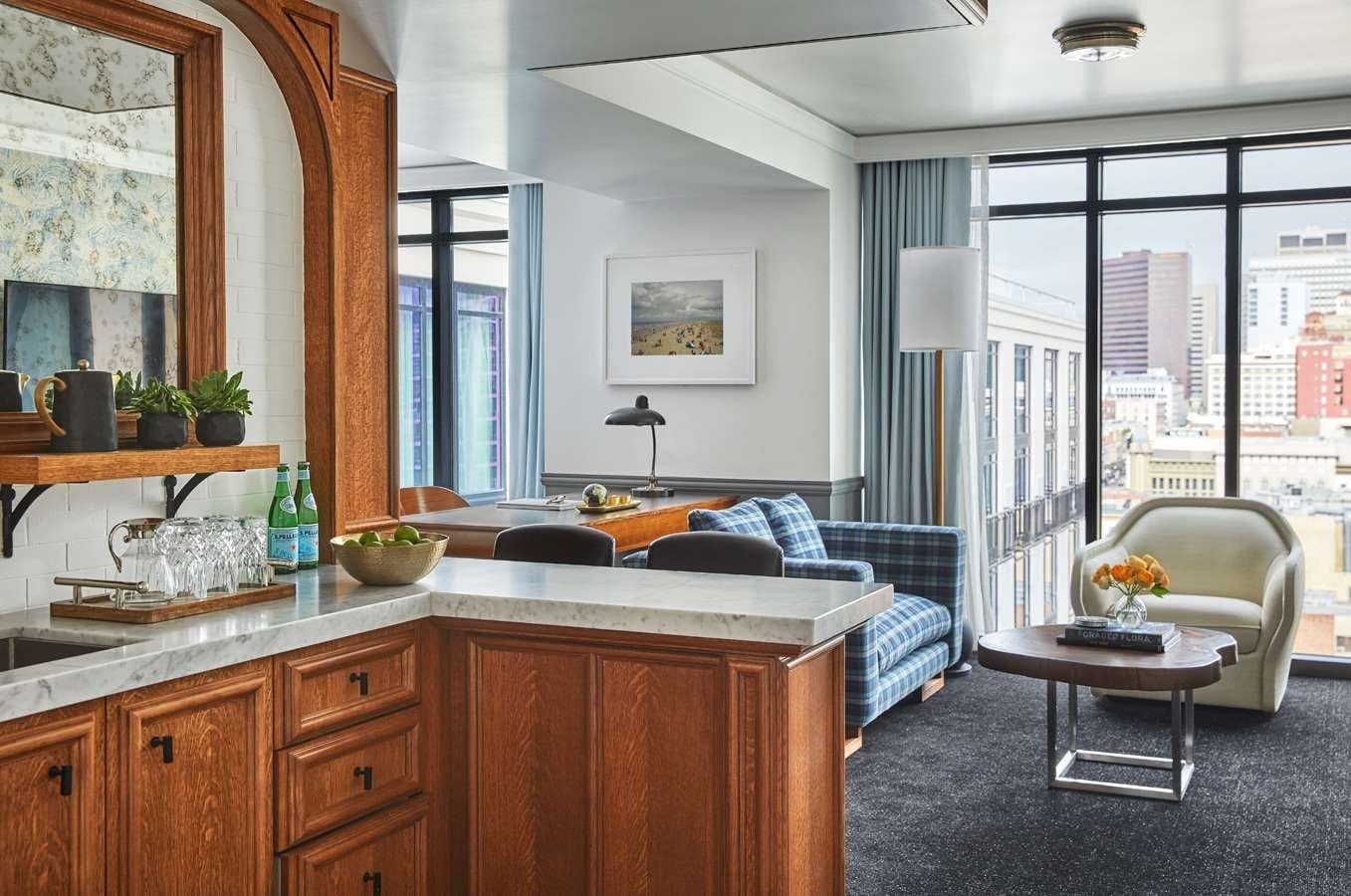 Une chambre d’hôtel moderne dotée d’un coin cuisine, d’un espace salon avec un canapé et des fauteuils, d’une grande fenêtre offrant une vue sur la ville et d’une petite table avec un vase.