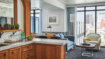 Une chambre d’hôtel moderne dotée d’un coin cuisine, d’un espace salon avec un canapé et des fauteuils, d’une grande fenêtre offrant une vue sur la ville et d’une petite table avec un vase.