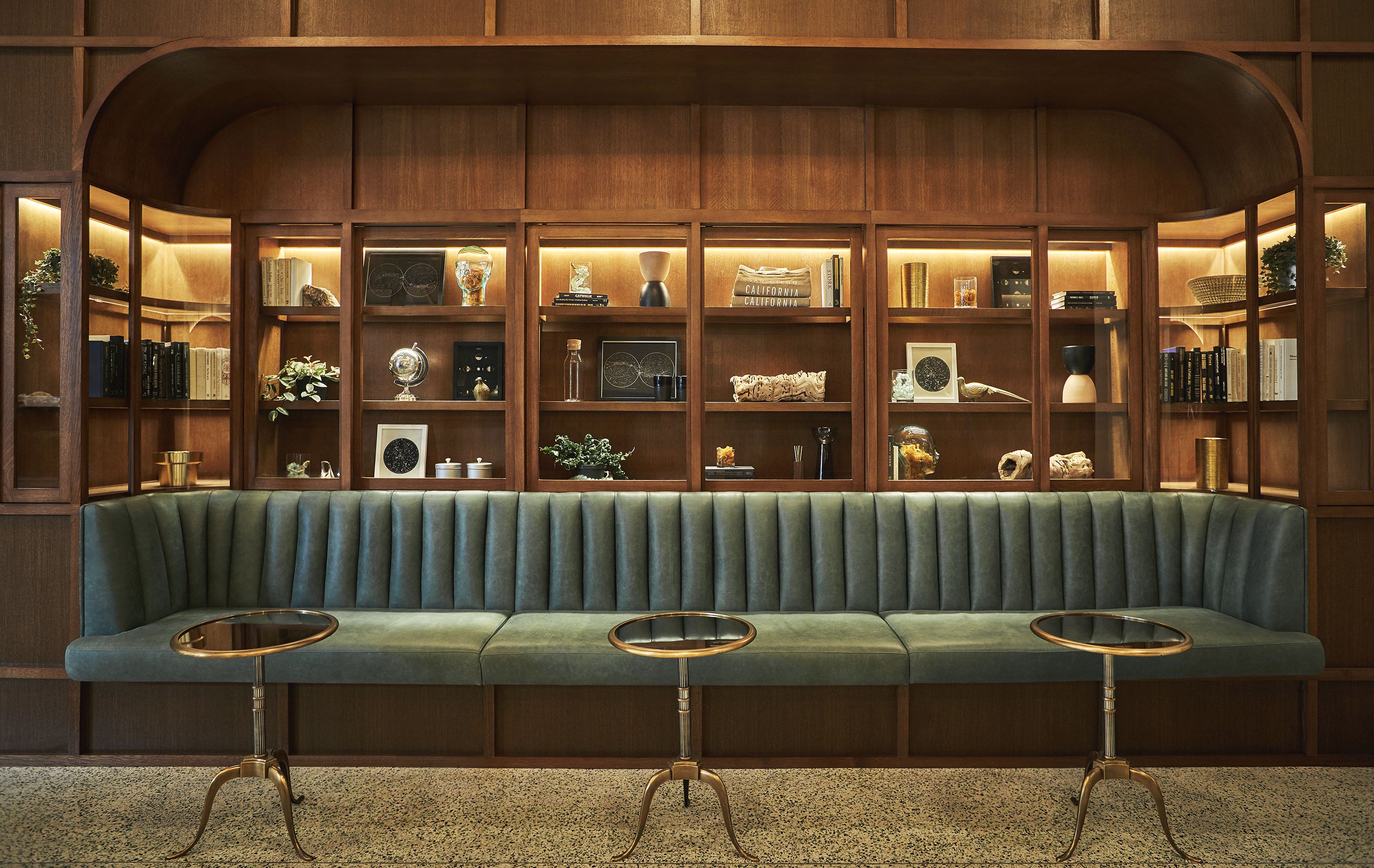 A green upholstered bench with two glass-topped tables in front, surrounded by wooden shelving with various items.