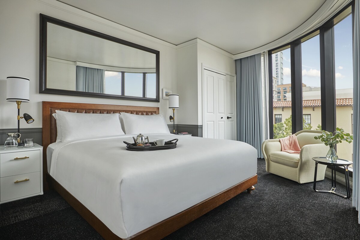 A hotel room with a large bed, a bedside table with a lamp, a chair, and a view of the city through large windows.