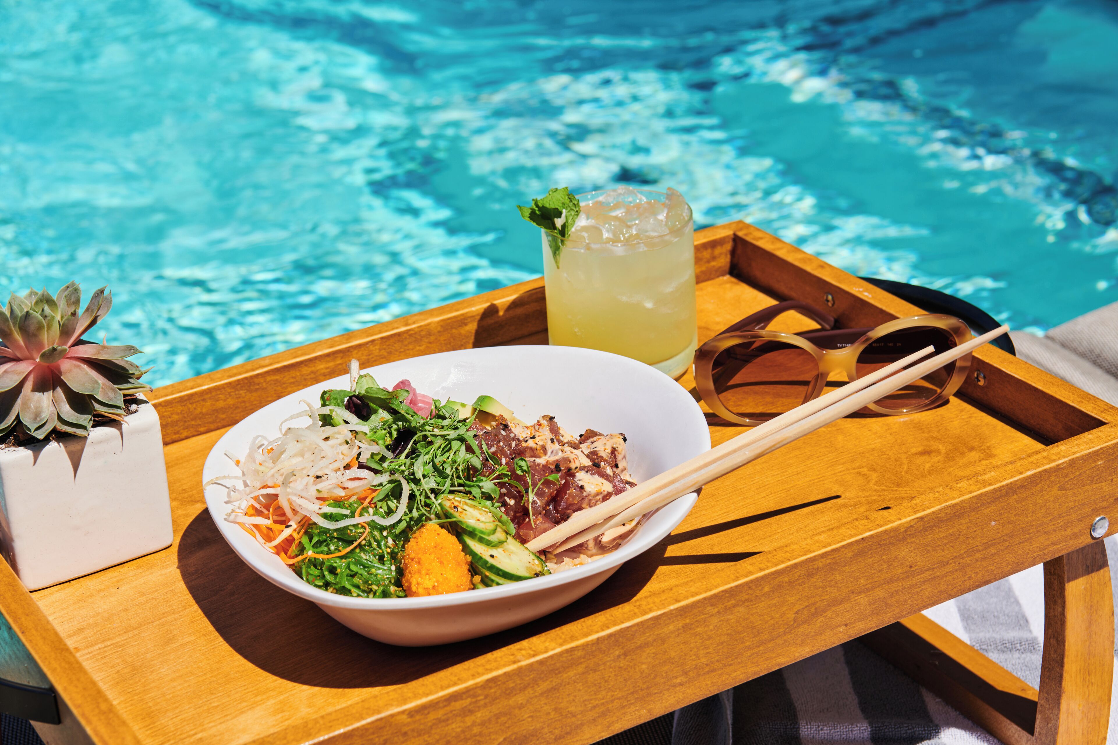 Una bandeja de madera con una ensalada, una bebida y palillos junto a una piscina.