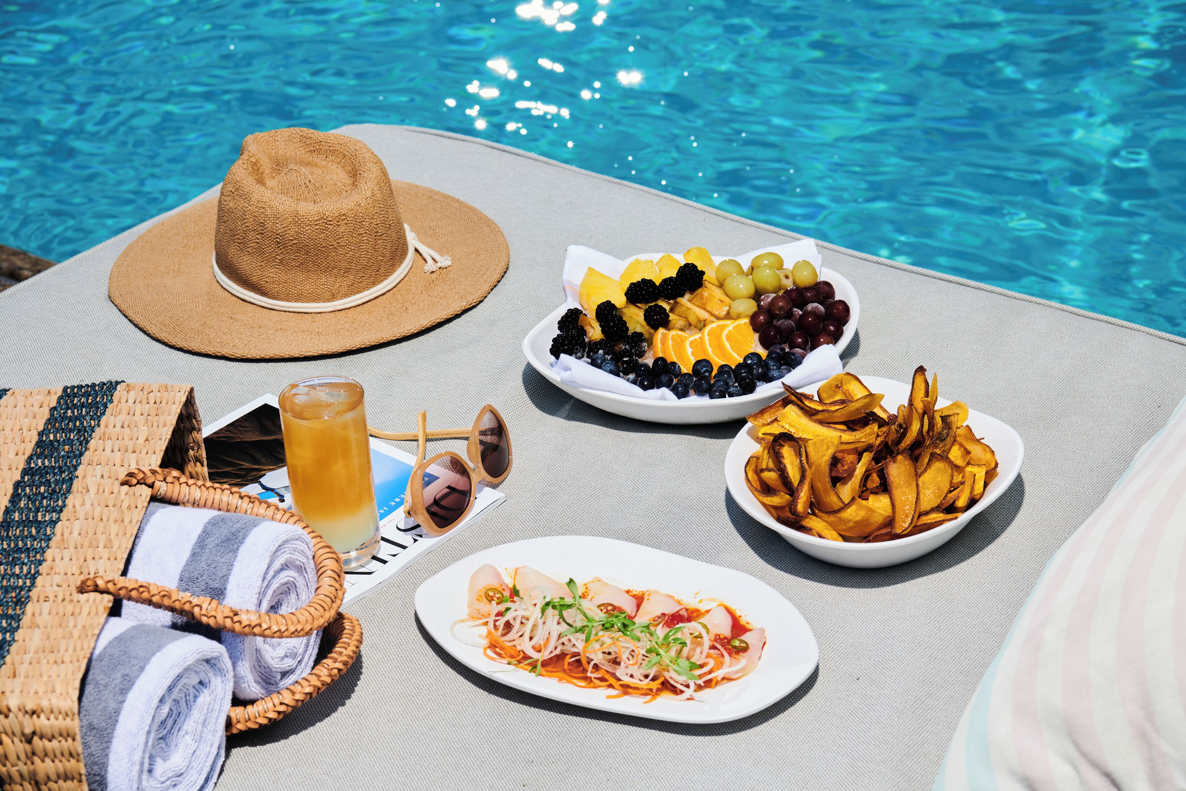 A poolside table with a hat, sunglasses, a basket, and plates of fruit and food.