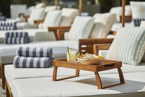 A row of sun loungers with white cushions and striped pillows, a wooden tray with a drink and snacks, and a view of the sea in the background.