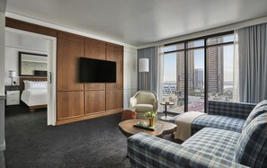 A modern hotel room with a large window, a flat-screen TV, a sofa, a chair, and a small table with a plant.