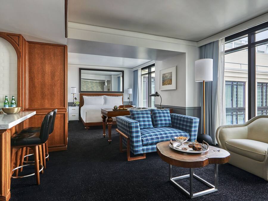 A hotel room with a bed, a blue plaid sofa, a small round table with snacks, a chair, a desk with a mirror, and a large window with curtains.