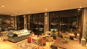 A modern living room with a grey sofa, wooden dining table, and chairs. There is a large window with a city view. - Bay View Guest House Somerset West (Cape Town)