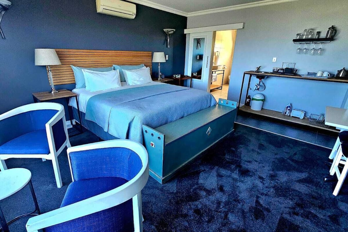 A hotel room with a bed, two chairs, a small table, a desk, and a kitchenette area.