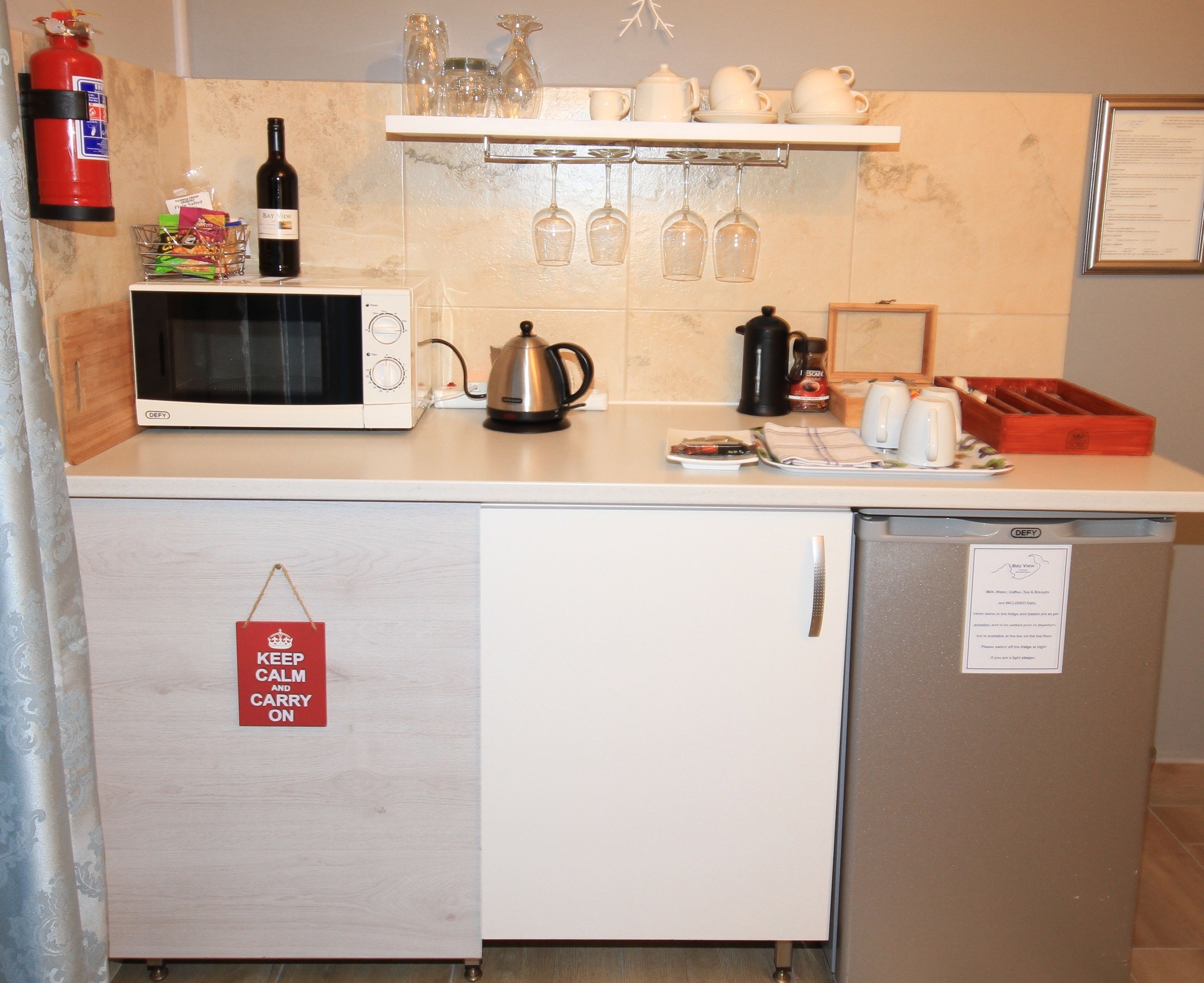 A kitchen with a microwave, kettle, and wine bottle. There is a fire extinguisher on the wall and a sign that says "Keep Calm and Carry On".