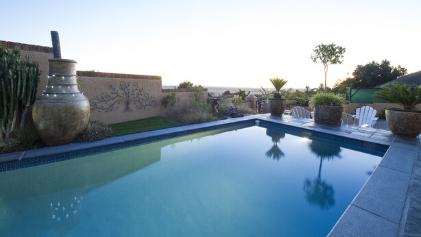 A swimming pool with a clear view of the surrounding landscape, including a cactus, a decorative wall, and a patio area with chairs and a table. - Bay View Guest House Somerset West (Cape Town)