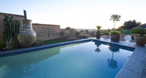 A swimming pool with a clear view of the surrounding landscape, including a cactus, a decorative wall, and a patio area with chairs and a table. - Bay View Guest House Somerset West (Cape Town)
