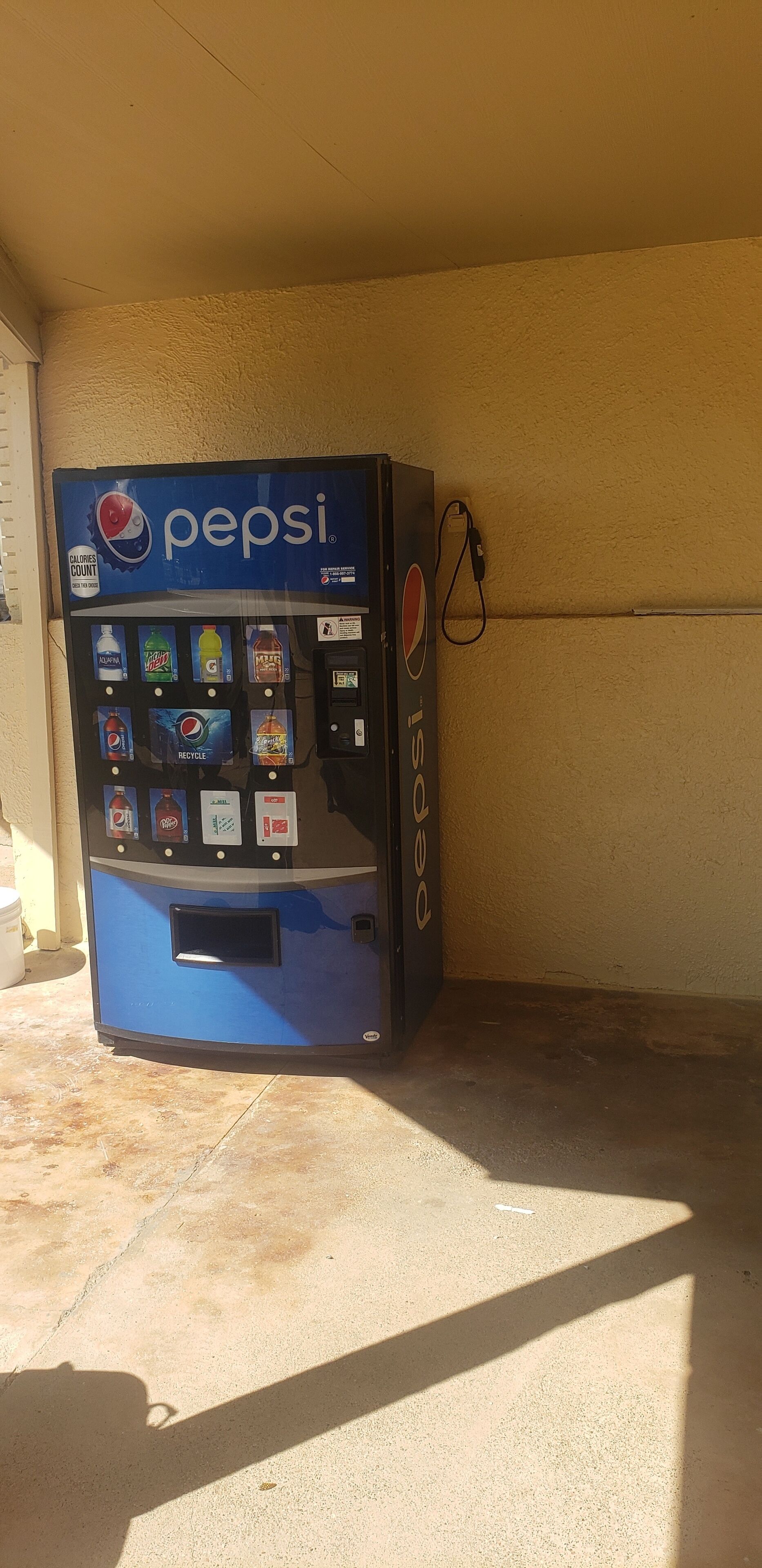 A Pepsi vending machine in a shaded area with a concrete floor.