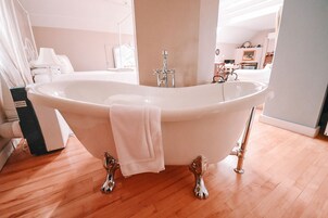 Una bañera independiente en una habitación con suelo de madera, una cama blanca y una mesita de noche.