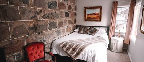 A bedroom with a stone wall, a bed with patterned pillows, a red chair, a bedside table with a lamp, and a painting on the wall.