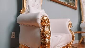 A luxurious white and gold upholstered chair with a tufted backrest and ornate legs, positioned in a room with a light blue wall and a framed landscape painting.
