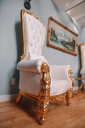 A luxurious white and gold upholstered chair with a tufted backrest and ornate legs, positioned in a room with a light blue wall and a framed landscape painting. - Bella's Castle (Morden)