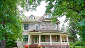 Une grande maison en pierre à deux étages, dotée d’une véranda qui fait le tour de la maison et d’un jardin bien entretenu.
