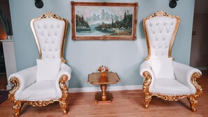 Deux fauteuils blancs richement décorés avec des motifs dorés, un tableau encadré représentant un paysage montagneux et une petite table en bois sur laquelle est posé un vase de fleurs.