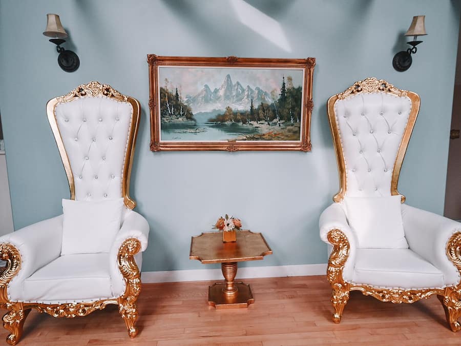Dos sillas blancas ornamentadas con detalles dorados, un cuadro enmarco de un paisaje montañoso y una pequeña mesa de madera con un florero.