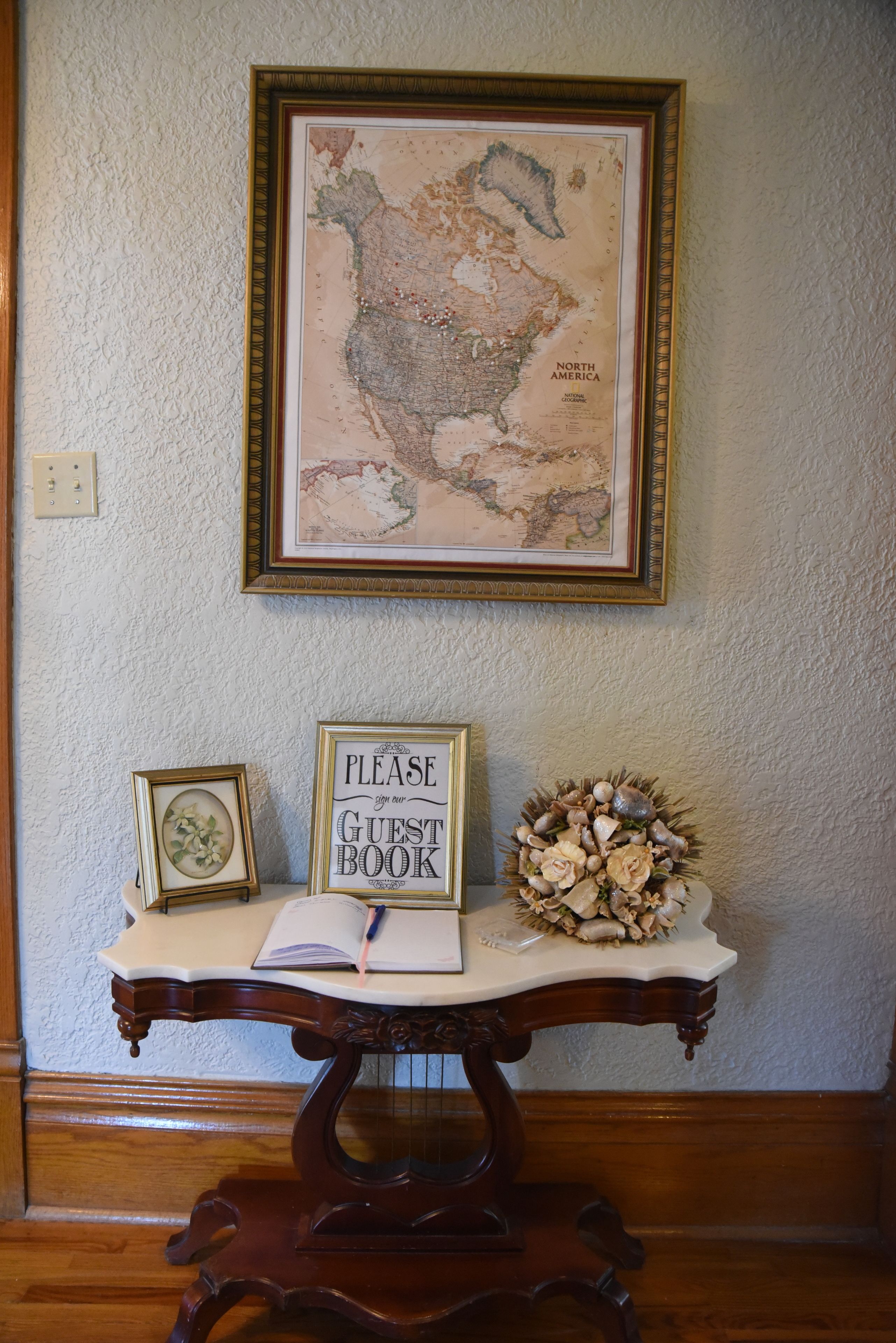 Un mapa enmarcado de Norteamérica en una pared, un letrero que dice “Por favor, firme su libro de visitas” sobre una mesa, y un arreglo floral decorativo.