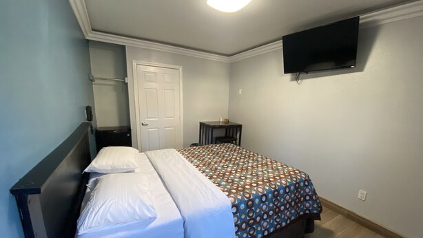 A bedroom with a bed, a bedside table, a chair, a desk, a TV mounted on the wall, and a door. - Sands Inn (South Gate)