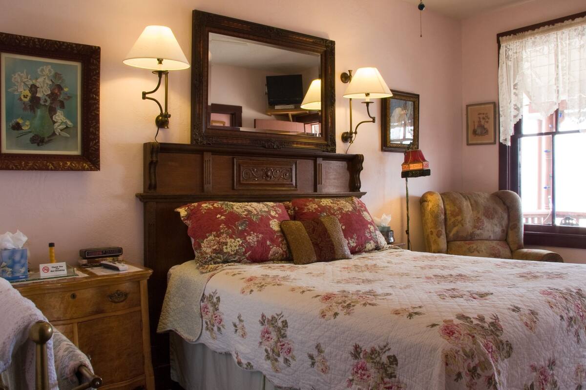 A bedroom with a large bed, two wall-mounted lamps, a wooden headboard, a chair, and a window with lace curtains.