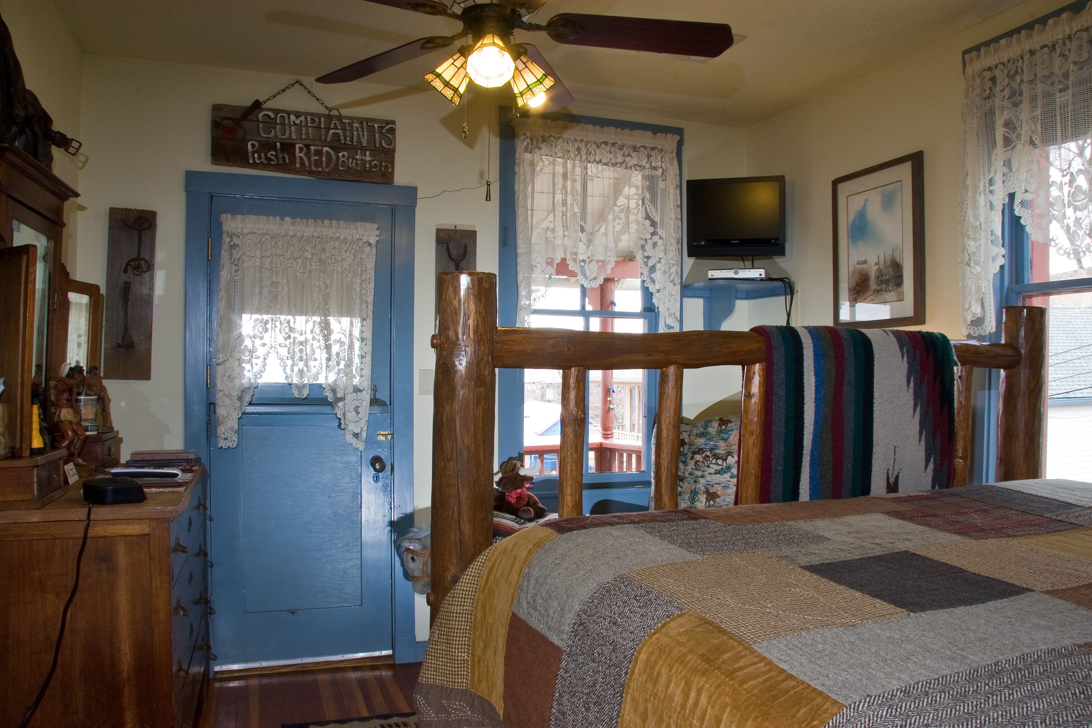 Un dormitorio con cama de madera, puerta azul, cortinas de encaje, ventilador de techo y televisor sobre un mueble bajo.