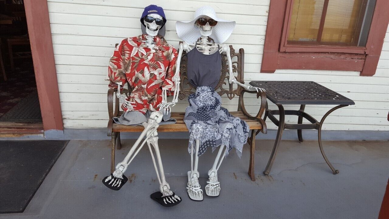 Dos figuras esqueléticas sentadas en un banco frente a un edificio con una ventana y una pequeña mesa cerca.