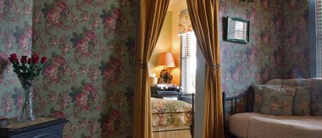 A room with floral wallpaper, a bed, a sofa, a small table with a vase of flowers, and a fireplace.