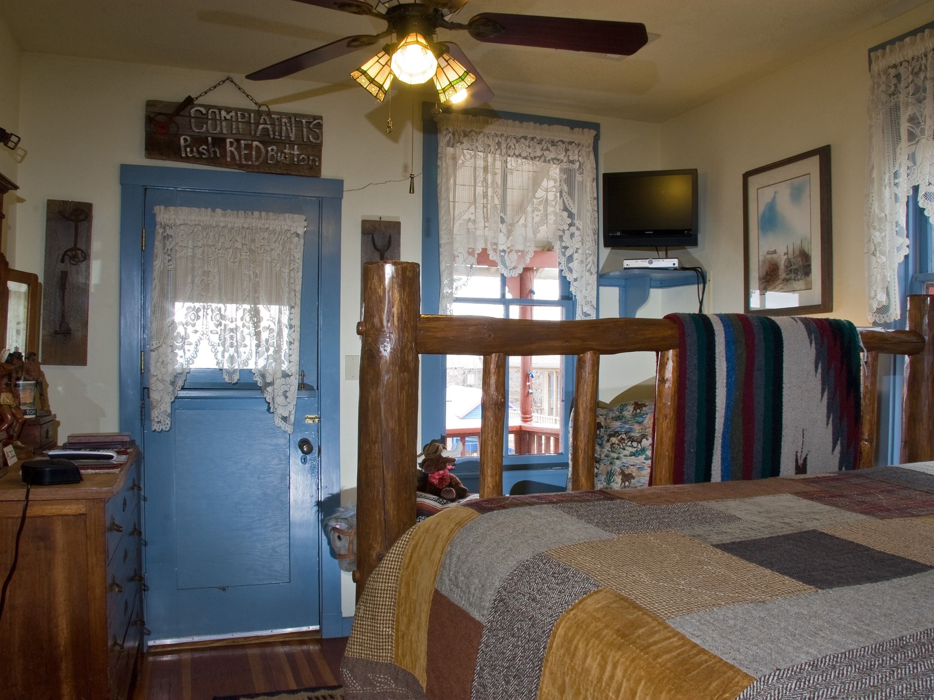 Un dormitorio con una cama de madera, una puerta azul, un ventilador de techo y un televisor en la pared.