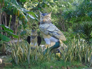 Deux grandes sculptures de hiboux, installées dans un jardin luxuriant, entourées d’un feuillage dense et de bananiers.