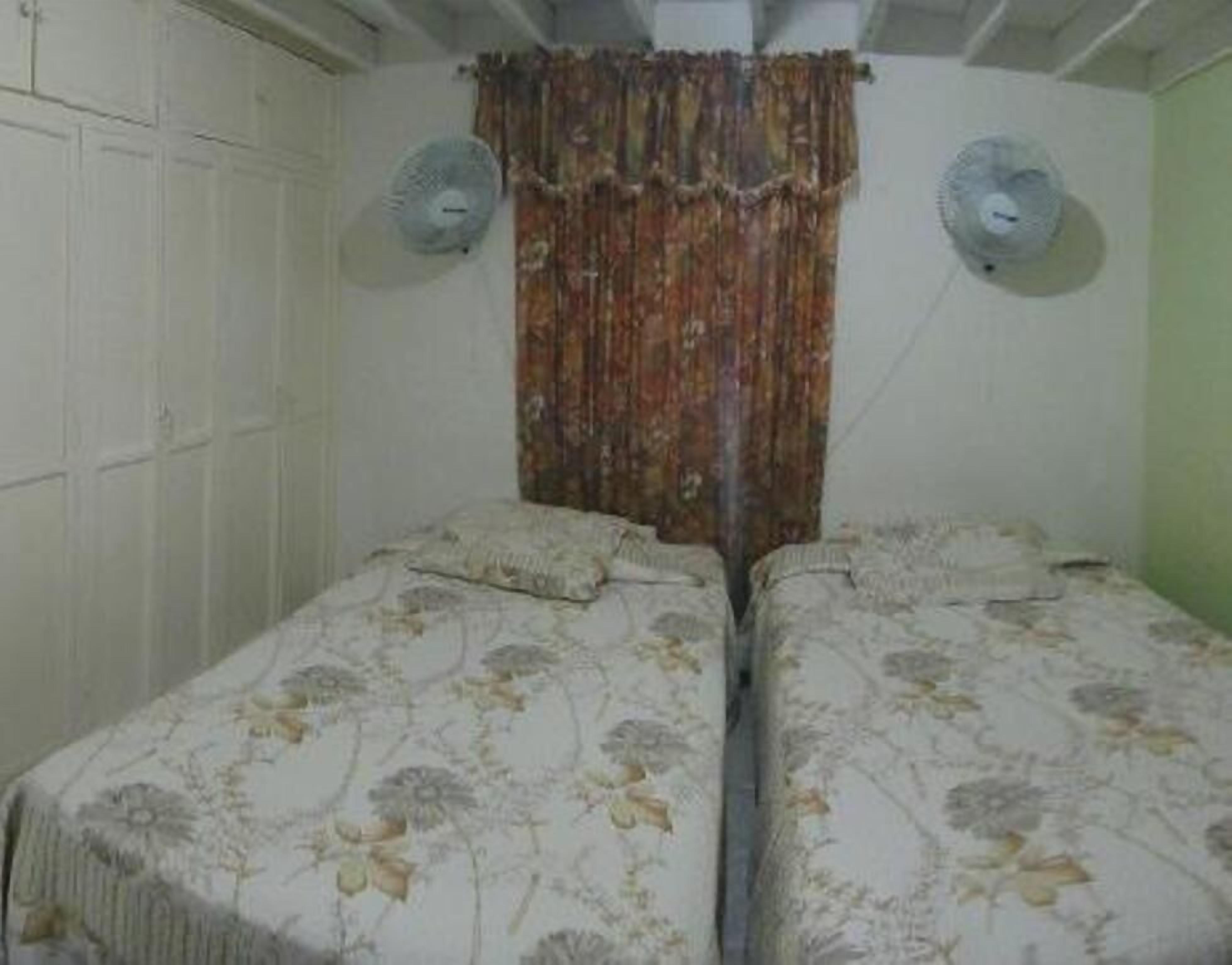 A room with two single beds, a patterned bedspread, a curtain, and two wall fans.