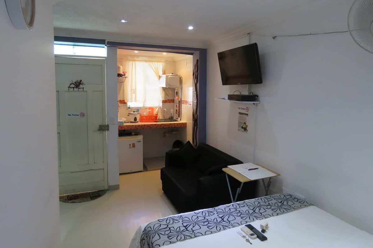 A compact room with a bed, a small table, a TV mounted on the wall, and a kitchenette in the background.
